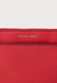 Červená kožená peněženka s texturovaným povrchem, zlatým logem "MICHAEL KORS" a zipovým uzávěrem podél horního okraje. Hladká vnitřní podšívka.