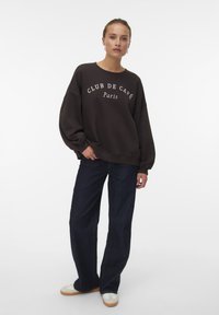 Vrouw in een grote donkere sweatshirt met de tekst "CLUB DE CAFÉ Parijs", donkere jeans en witte sneakers, staande met één hand in de zak.