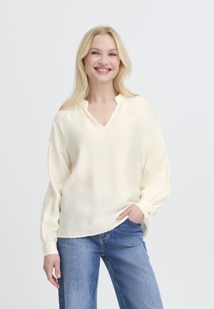 Donna sorridente con lunghi capelli biondi, indossa una camicetta cremosa e ampia e jeans blu, in piedi con una mano in tasca.