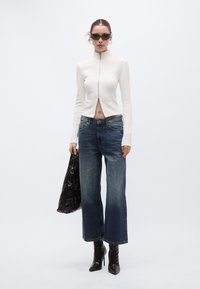 Λευκό κοντό μπλουζάκι με φερμουάρ μπροστά, συνδυασμένο με μπλε τζιν παντελόνια φαρδιάς γραμμής και μαύρες μπότες με τακούνι. Κρατάει μια μαύρη τσάντα.