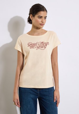 T-shirt en coton couleur crème à manches courtes, arborant du texte en rouge en cursive "De bonnes choses arrivent" centré sur le devant.