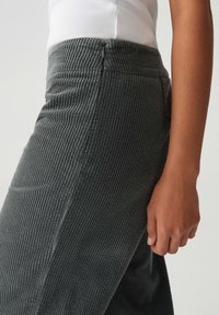 Gros plan sur une personne portant un pantalon à côtes gris foncé avec une fermeture éclair sur le côté et un débardeur à côtes blanc, le bras détendu le long du corps.
