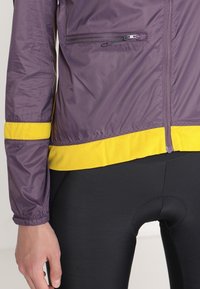 Lätt lila jacka med gula detaljer vid fållen och ärmsluten. Har en zip-ficka på sidan, tillverkad av lätt och ventilerande tyg.