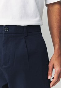 Granatowe spodnie o eleganckim kroju, z zapięciem na guzik, przednimi zakładkami i gładką fakturą tkaniny. Częściowo widoczna ręka z boku.