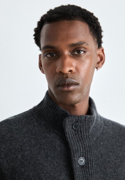 Pull en tricot gris foncé avec un col montant et une texture côtelée, comportant deux boutons noirs au niveau du cou. Gros plan sur le vêtement.