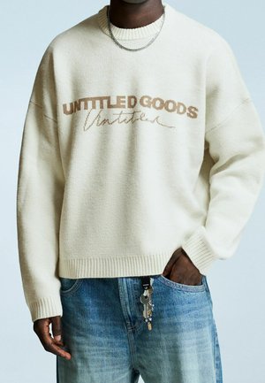 Personne portant un pull beige avec le texte "UNTITLED GOODS" et un jean bleu, main dans la poche tenant des clés attachées à la boucle de ceinture.