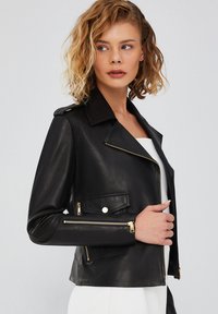 Zwarte leren jas met een getailleerde pasvorm, ritsmouwen, gouden hardware accenten en een gekartelde kraag. Bevat een detail met een zak aan de voorkant.