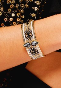 Large bracelet métallique présentant un motif de perles en or, noir et argent, avec deux pierres précieuses grises en forme de goutte au centre.