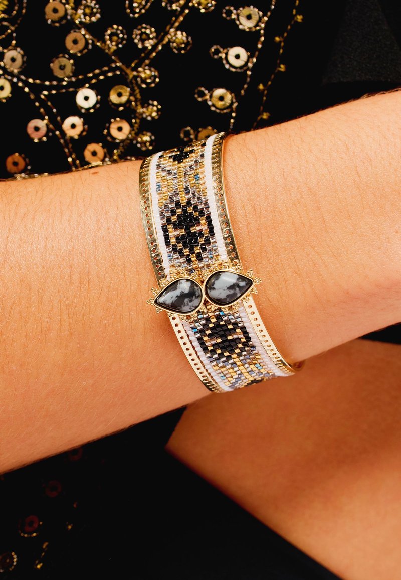 Large bracelet métallique présentant un motif de perles en or, noir et argent, avec deux pierres précieuses grises en forme de goutte au centre.