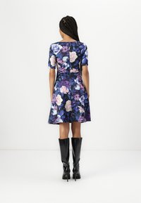 Virágos ruha sötétkék háttérrel, rövid ujjakkal, testhez simuló felsőrésszel és bővülő szoknyával; fekete térd fölé érő csizmákkal párosítva.