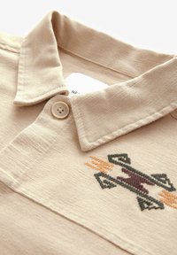 Beige katoenen polo shirt met een kraag, voorzien van een knoopsluiting en ingewikkelde borduursels in groen, bruin en oranje.