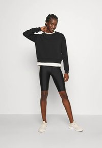 Sudadera negra recortada con cuello redondo, combinada con pantalones cortos de ciclista negros brillantes. La modelo está de pie con zapatillas deportivas neutras, sonriendo, sobre un fondo claro.