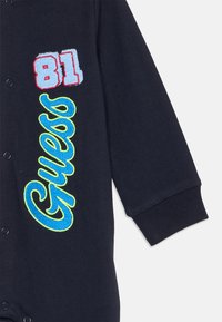Body en coton marine avec broderie rouge et bleue "81" et broderie verte et bleue "Guess," doté de fermetures à pression et de poignets côtelés.