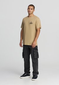T-shirt di cotone beige con maniche corte e un logo sul petto, abbinata a pantaloni cargo neri con tasche laterali e una vestibilità rilassata.