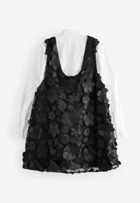 Schwarzes, floral gemustertes, ärmelloses Kleid mit tiefem Rücken, über einem weißen kragenlosen Hemd getragen. Strukturiertes Gewebe mit 3D-Blumenakzenten.