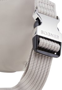 Vaalea beige ristikkäinen olkalaukku, jossa on teksturoitu hihna ja hopeinen metallisolki, johon on kaiverrettu "BOGNER". Kestävä materiaali, minimalistinen muotoilu.