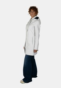 Leggera giacca a vento bianca con cappuccio, cerniera frontale e tasche laterali, abbinata a jeans svasati scuri e sneakers beige.