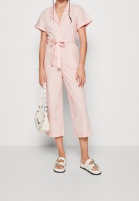 Light pink belted jumpsuit with short sleeves, wide cropped legs, and two front pockets. Paired with white sandals and a cream handbag.