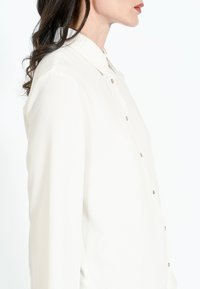 Biała koszula z guzikami, klasycznym kołnierzykiem, długimi rękawami i delikatnym połyskiem. Posiada metalowe guziki z przodu do zapinania.