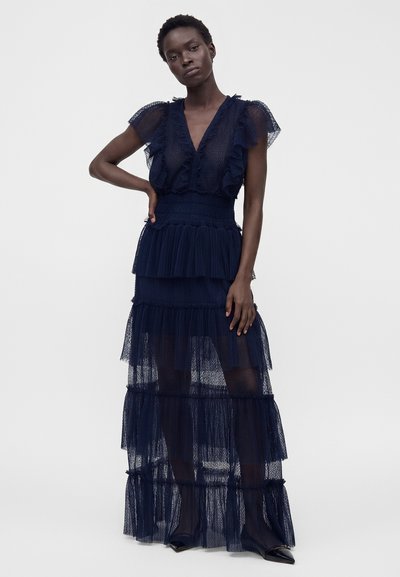 Femme aux cheveux courts et bouclés portant une robe à étages bleu marine transparente avec des manches volantées et des chaussures noires pointues, debout, la main sur la hanche.