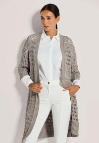 Langer, offen gestrickter Cardigan in Hellgrau, mit einer strukturierten Webart, gerippten Bündchen und einer lockeren Form. Kombiniert mit einem weißen Hemd und Hose.