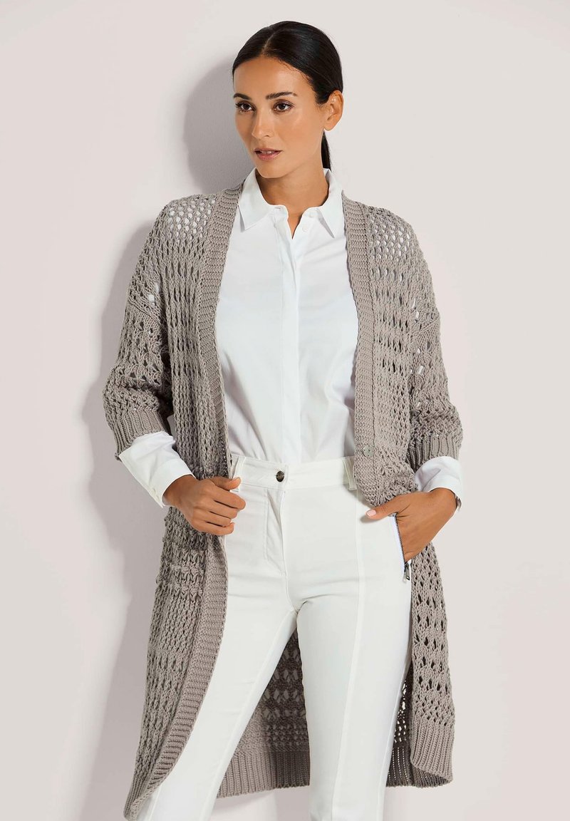 Langer, offen gestrickter Cardigan in Hellgrau, mit einer strukturierten Webart, gerippten Bündchen und einer lockeren Form. Kombiniert mit einem weißen Hemd und Hose.