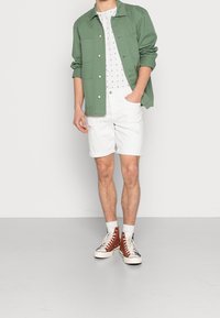 Chaqueta verde de botones sobre una camisa blanca con pequeños estampados de veleros en navy, combinada con pantalones cortos blancos y zapatillas altas marrones.