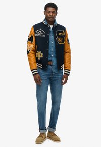 Chaqueta de béisbol azul marino con mangas de cuero naranja, parches bordados, camisa de mezclilla negra, jeans azules y zapatos color marrón claro.