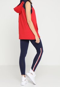 Kvinde iført en ærmeløs rød hoodie, navy leggings med rød og hvid stribe samt hvide sneakers, som står foran en hvid baggrund.