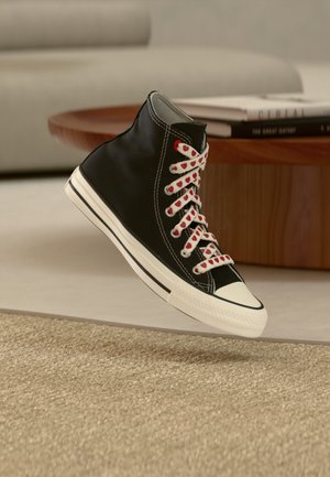 Baskets montantes noires avec semelle blanche et lacets rouge à motifs de cœurs flottant au-dessus d'un tapis beige dans un salon.