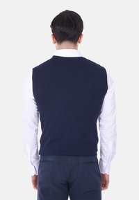 Maglione senza maniche blu navy, texture a maglia, scollo rotondo, indossato sopra una camicia bianca a maniche lunghe, visto da dietro.