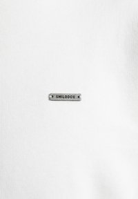 T-shirt in cotone bianco con una piccola etichetta in metallo grigio impresso con "SMILODOX" attaccata vicino al colletto. Texture liscia, design minimale.