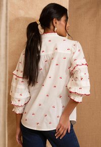 Blusa bianca con ricamo di ciliegie rosse, maniche a volant e scollo rotondo. Tessuto morbido con silhouette rilassata e orlo curvo.