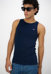 Tricou fără mâneci de culoare bleu marin pentru bărbați, fabricat dintr-un material elastic, cu guler rotund și un mic logo al brandului pe piept. Purtați cu ochelari de soare închiși la culoare.
