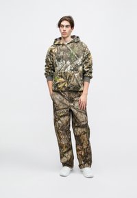 Σετ με καμουφλαρισμένο φούτερ και παντελόνι με σχέδιο φύλλων και κλαδιών σε γήινες αποχρώσεις, με μπροστινή τσέπη και ελαστικούς καρπούς, συνδυασμένο με λευκά αθλητικά παπούτσια.