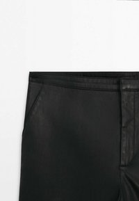 Section de la taille d'un pantalon en cuir noir montrant les passants de ceinture, la poche latérale et la couture avant sur fond blanc.