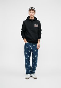 Musta huppari, jossa on etutasku ja logopatch, yhdistettynä sinisiin farkkuihin, joissa on toistuvia yin-yang-kuvioita. Valkoiset tennarit.