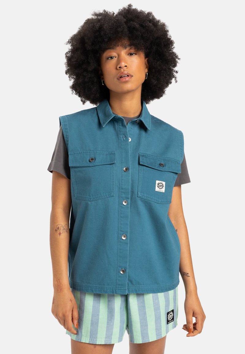 Tealblauwe mouwloze shirt met twee borstzakken, knopen en een wit logo label. Model draagt gestreepte groene en blauwe shorts.