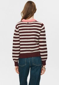 Frau mit langen Haaren, die einen weinroten und weißen gestreiften Pullover mit rosa Kragen und blauen Jeans trägt, von hinten betrachtet.