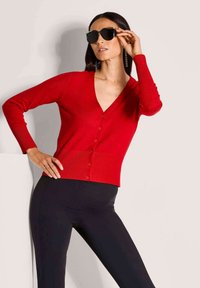 Roter Strickcardigan mit langen Ärmeln, tiefem V-Ausschnitt und Knopfverschluss. Kombiniert mit schwarzen Leggings und schwarzen Sonnenbrillen.