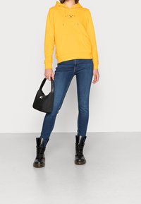 Gelber Hoodie mit Logo, blaue Skinny-Jeans, schwarze Knöchelstiefel und eine kleine schwarze Tasche mit Logoschlaufe. Einfaches, lässiges Ensemble mit einem einheitlichen Farbschema.
