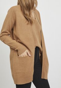 Chaleco de punto marrón con un corte holgado, puños acanalados y detalles en los bolsillos. Blusa de escote en V debajo, combinada con pantalones negros.