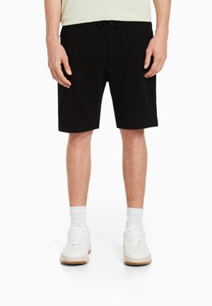 Czarne bawełniane szorty z elastycznym paskiem i sznurkiem do wiązania. Gładka faktura, fason prosty, zestawione z białymi trampkami i skarpetkami.