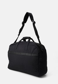 Sac de sport noir en tissu durable, doté de doubles poignées supérieures, d'une bandoulière amovible et d'une fermeture zippée avec une poche plate.