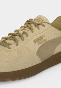Κοντινή λήψη μπεζ αθλητικού παπουτσιού Puma Palermo με υφή από σουέτ, κορδόνια με σχέδια και καφέ λαβή από καουτσούκ σε λευκό φόντο.