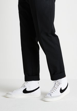 High-top sneakers with white leather, black suede Nike swoosh, gray toe cap, and black pants with rolled cuffs, standing on a light surface.