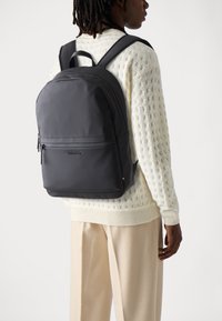 Zaino nero realizzato in materiale liscio, dotato di una tasca frontale con zip, spallacci imbottiti e un logo sul davanti.