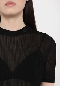 Czarny krótki top z prążkowaną fakturą, z subtelnym logo na kołnierzu oraz warstwowym projektem z półprzezroczystymi elementami.