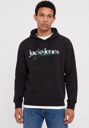 Jeune homme portant un sweat à capuche noir Jack & Jones avec des lettres florales, un jean bleu et une chemise blanche en dessous, debout sur fond blanc.