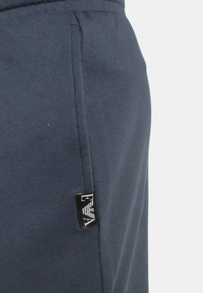 Tessuto blu navy con una superficie liscia, presenta una tasca laterale e una piccola etichetta nera con la scritta "EVA". Design a vestibilità sagomata.